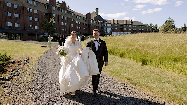A bride and groom in wedding attire stroll down a gravel path beside a grassy lawn and a lodge building during daylight.