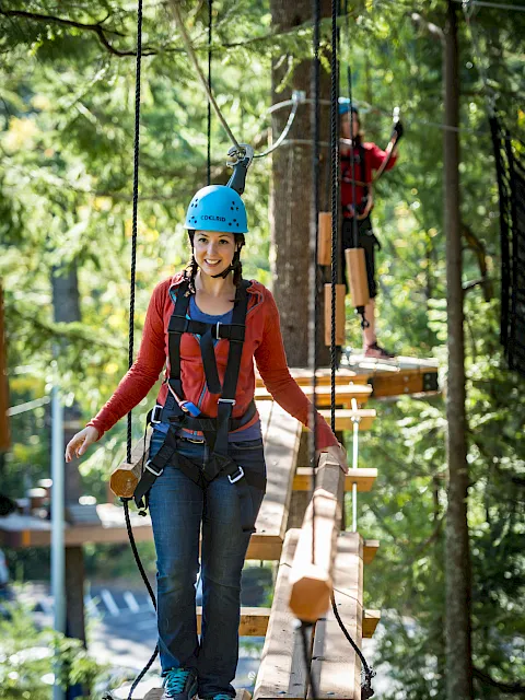 A woman in a blue helmet and safety harness stands on a rope bridge in a forest adventure park, smiling.
