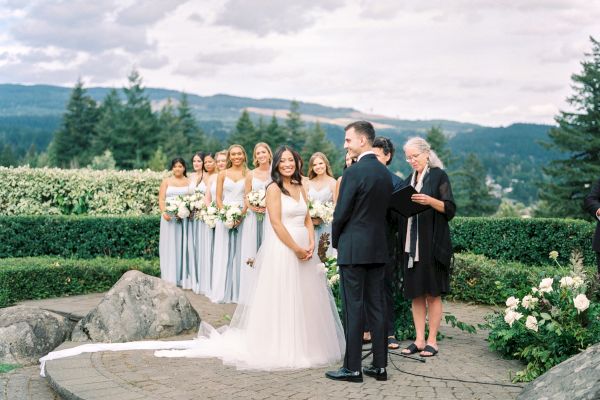 A wedding ceremony with a bride and groom standing together outdoors, bridesmaids holding bouquets, and an officiant present.