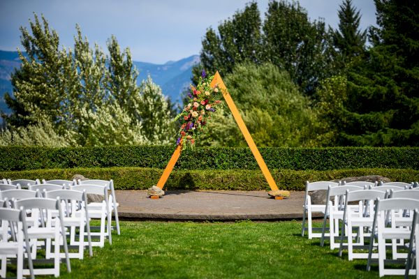 An outdoor wedding setup with an empty aisle, white chairs, and a triangular wooden arch adorned with flowers and greenery.