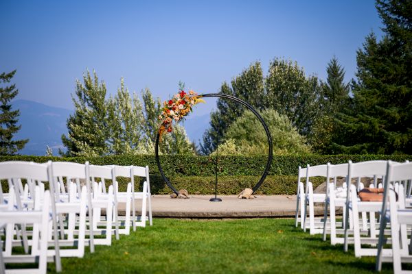An outdoor wedding setup with white chairs facing a decorative circular arch adorned with flowers, set against a backdrop of trees and clear skies.