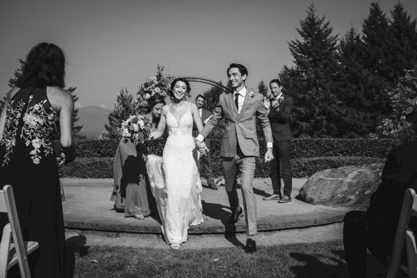 A newlywed couple walks down the aisle, smiling, outdoors with guests and a bridal party applauding.