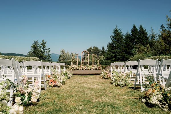The image shows an outdoor wedding setup with white chairs, a floral aisle, and an arch at the front, surrounded by greenery and blue skies.