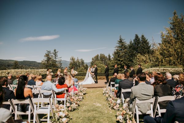 A wedding ceremony outdoors with guests seated and a couple at the altar surrounded by trees and greenery.