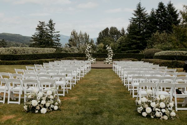 An outdoor wedding setup with white chairs and floral arrangements, set on a grassy area surrounded by trees and distant hills.