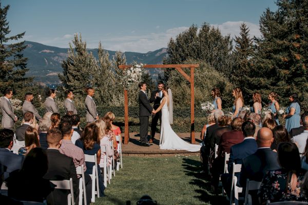 A couple is having an outdoor wedding ceremony with guests seated, surrounded by greenery and mountains in the background.