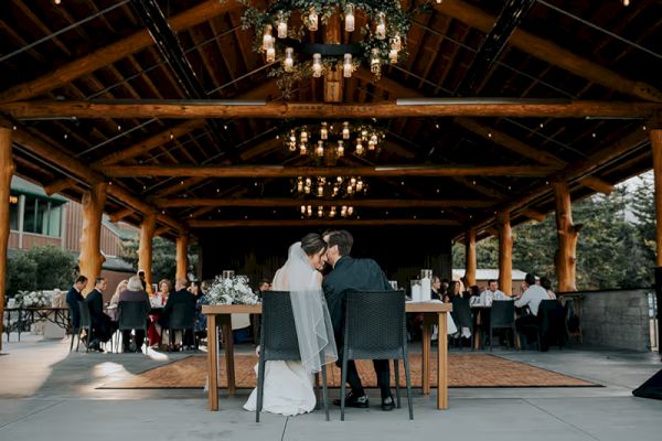 A couple in wedding attire sits at a table in a rustic venue with guests nearby, under a wooden ceiling with hanging lights.