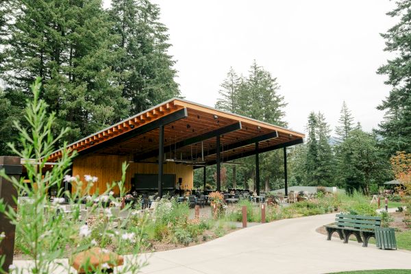 The image shows an outdoor pavilion with a wooden roof surrounded by trees, seating, and a garden-like setting.