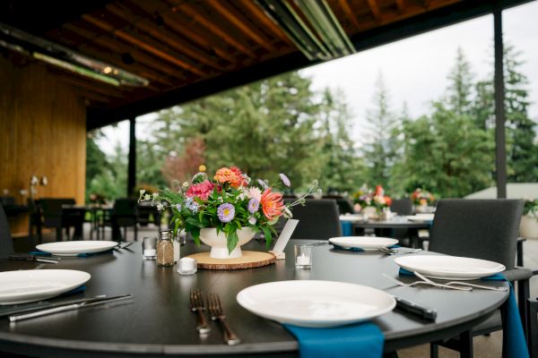An outdoor dining setup with a table adorned with a floral arrangement, plates, and cutlery, surrounded by trees under a wooden roof.