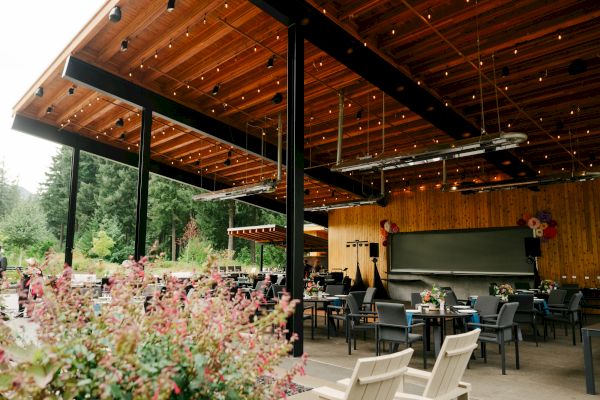An outdoor restaurant patio with wooden ceiling, string lights, and empty tables and chairs surrounded by greenery and plants.