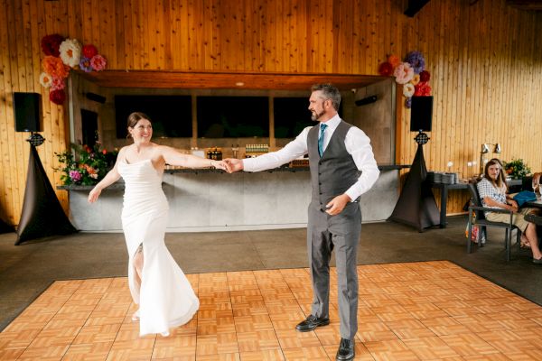 A couple is dancing on a wooden floor. The woman wears a white dress, and the man is in a suit. The setting is a decorated hall.