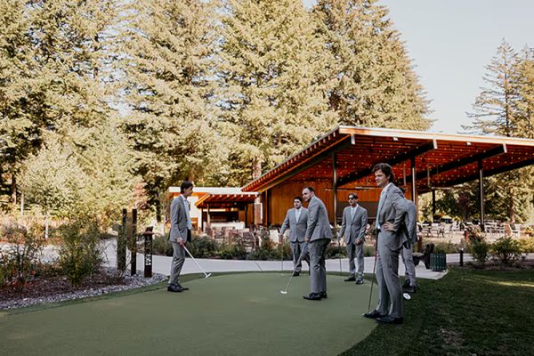 A group of people in suits stand on a putting green near a wooden building, surrounded by trees under a clear sky.
