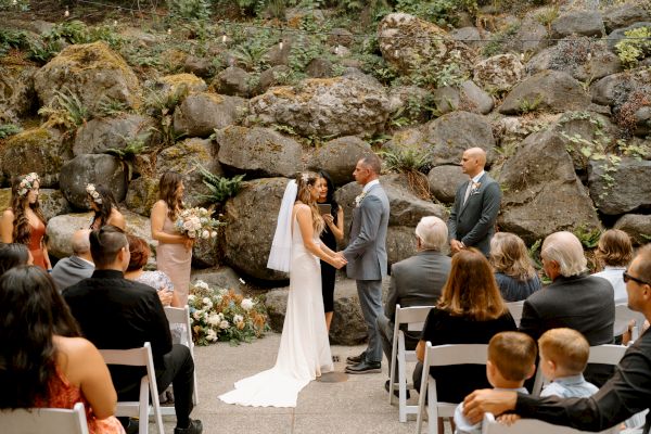 A wedding ceremony is taking place outdoors, with a couple exchanging vows in front of guests seated on white chairs.