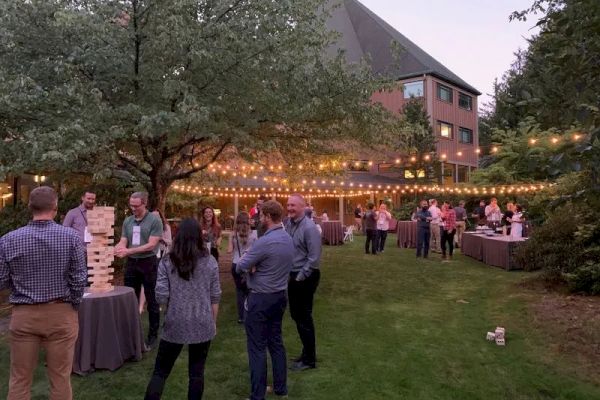 A group of people are enjoying an outdoor gathering, playing giant Jenga under string lights on a grassy area.