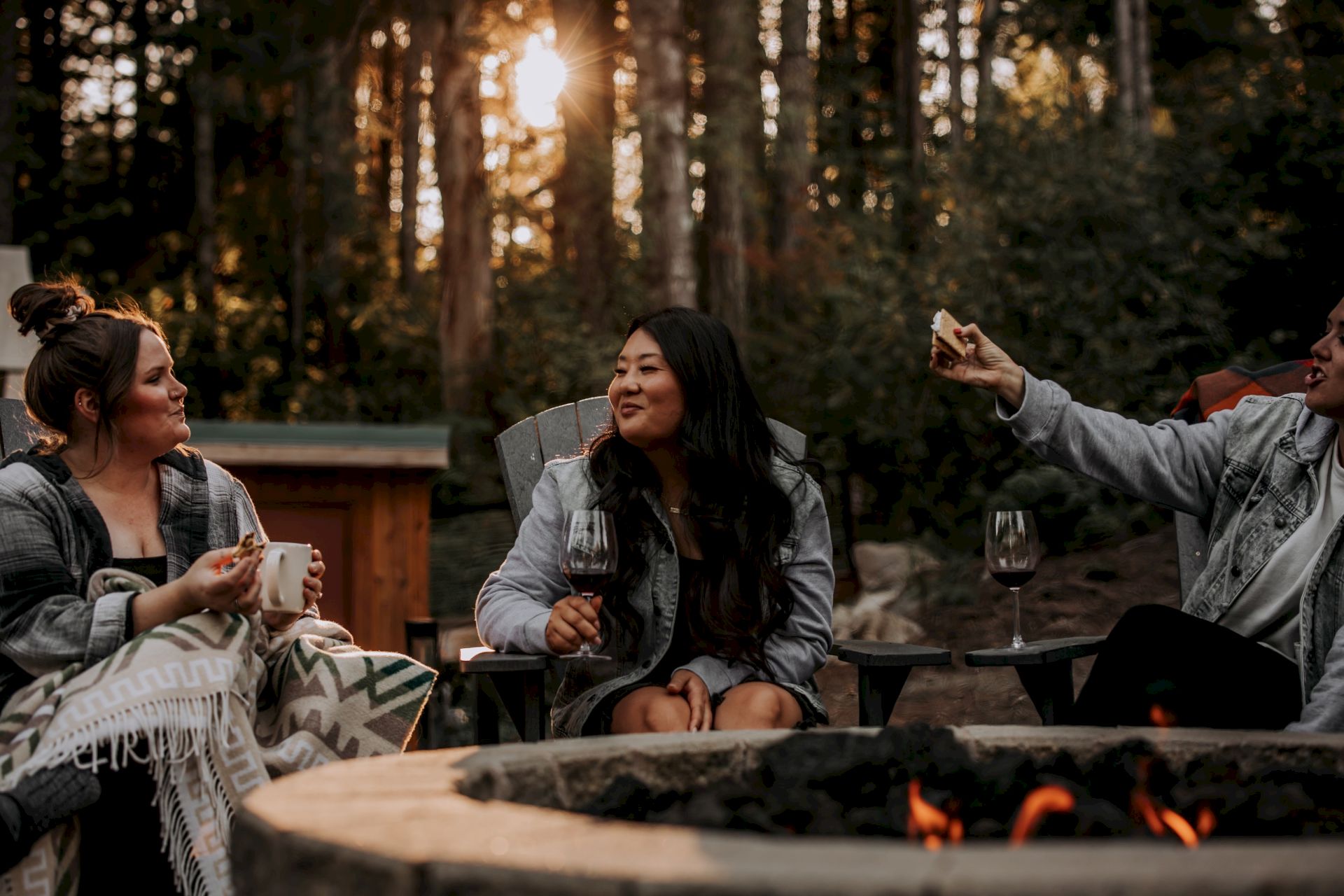 Three friends toast with wine by an outdoor firepit in a forest at sunset.