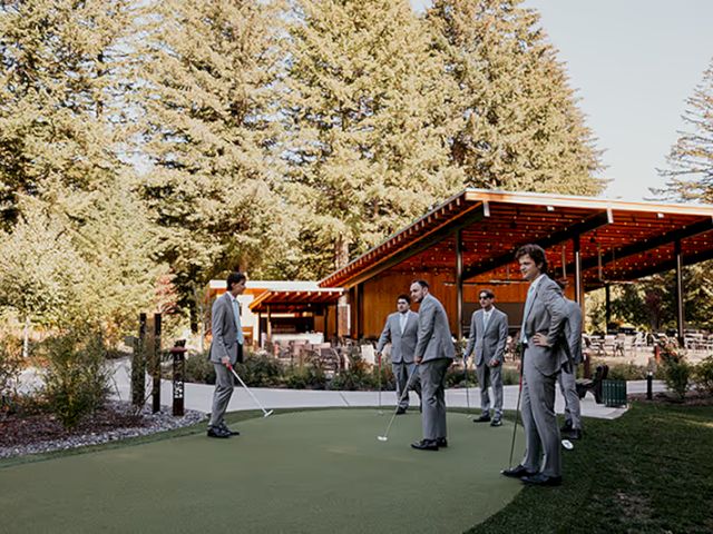 A group of people in suits are standing on a green outdoor area with golf clubs, near a wooden pavilion surrounded by trees.