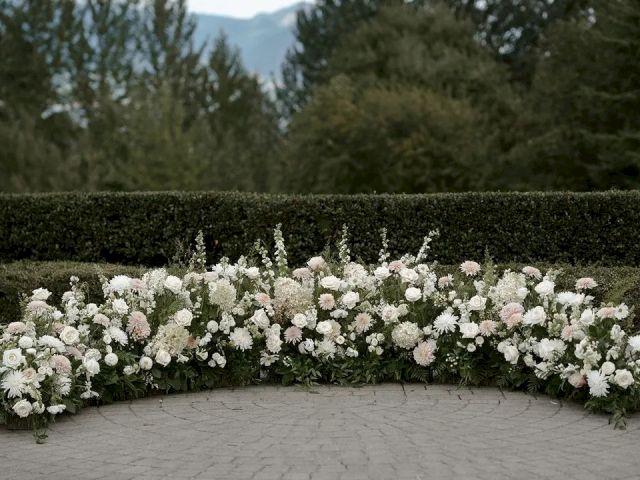 The image shows an arrangement of white and pink flowers on a pavement, with trees and hedges in the background, under a cloudy sky.