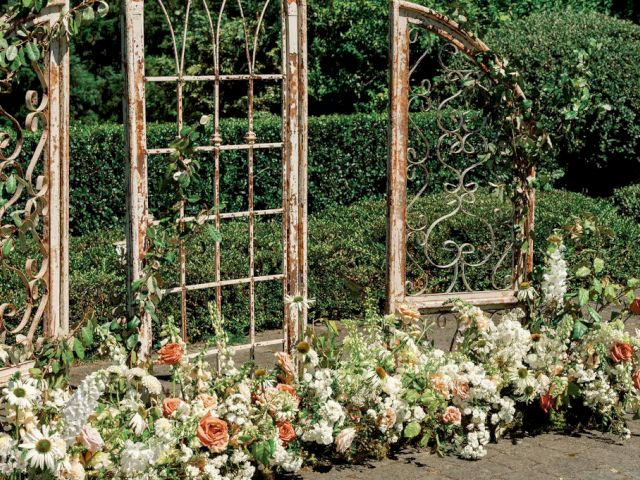 An ornate, rustic metal trellis setup adorned with climbing plants and surrounded by various flowers in a garden setting.