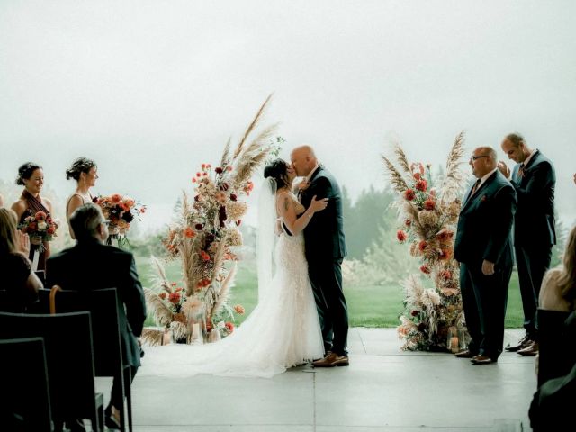 A couple is kissing at a wedding ceremony, surrounded by guests and floral arrangements, with an outdoor backdrop.
