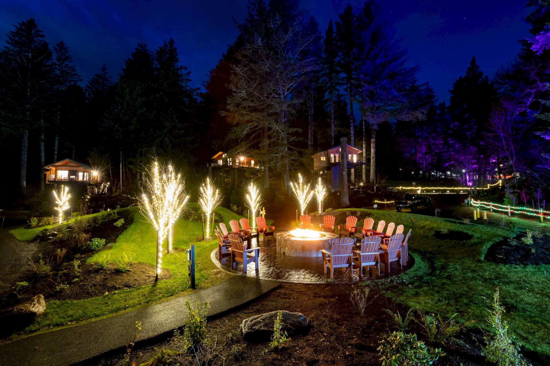 A nighttime forest lodge patio glows with colorful lights, chairs around a fire pit, and sparking fountains in the yard.