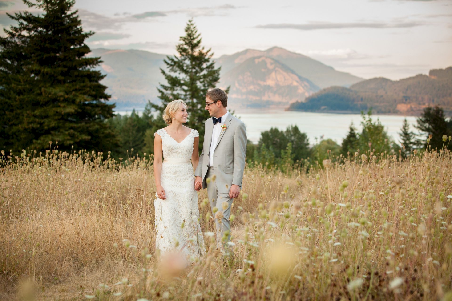 A bride and groom hold hands in a field with pine trees and mountains behind them, outdoors on their wedding day.