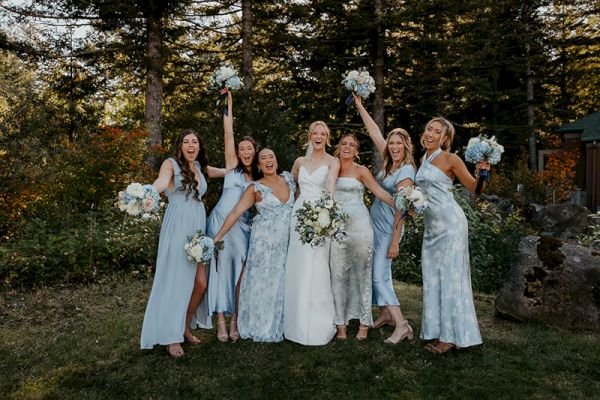 A group of women in dresses pose joyfully outdoors, holding bouquets, surrounded by trees and greenery, celebrating an event.