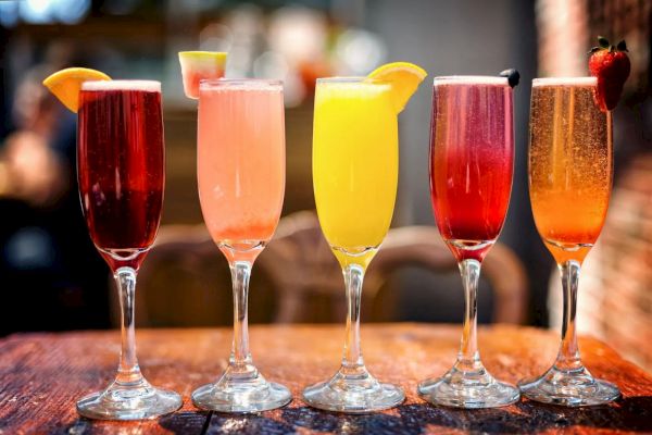 Five colorful cocktails in fluted glasses, each with different garnishes like fruits, arranged on a wooden table.