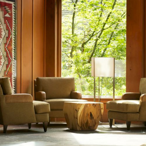 A cozy seating area with three armchairs, a wooden table, and a standing lamp. A decorative tapestry hangs on the wood-paneled wall.