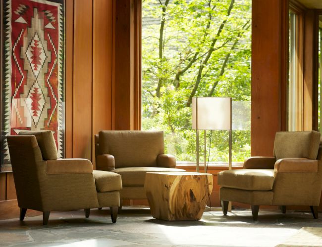 A cozy seating area with three armchairs, a wooden table, and a standing lamp. A decorative tapestry hangs on the wood-paneled wall.