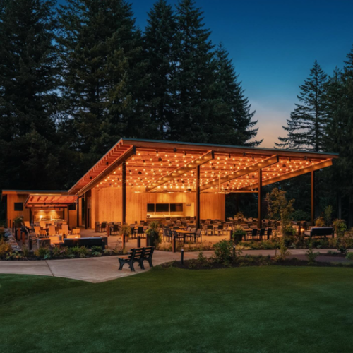 An outdoor pavilion with string lights, surrounded by trees, features seating and tables, creating a cozy evening atmosphere.
