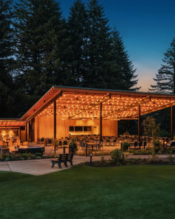 An outdoor pavilion with string lights, surrounded by trees, features seating and tables, creating a cozy evening atmosphere.