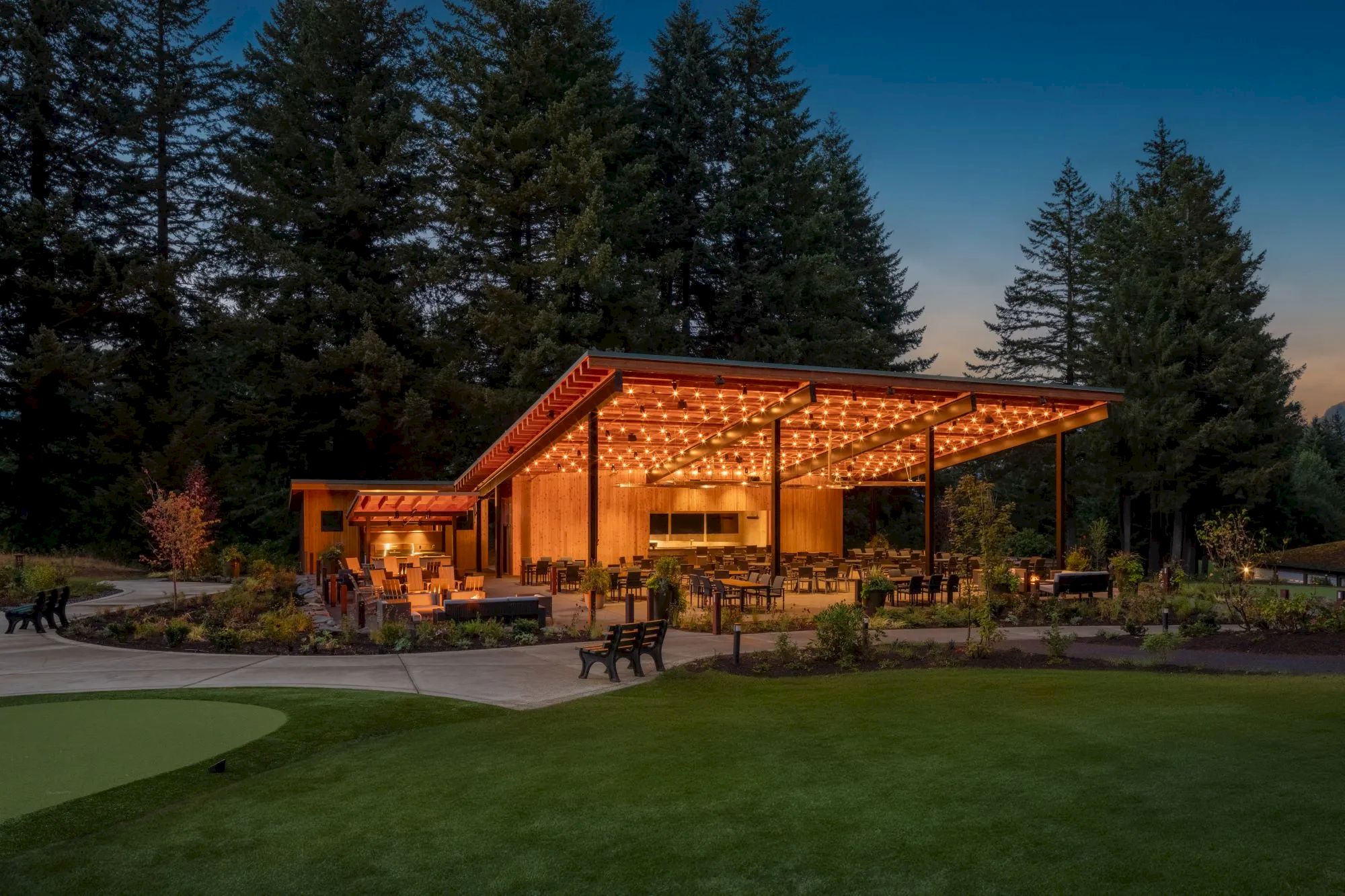 A modern outdoor pavilion with string lights, surrounded by trees and greenery, features seating and a bar area in a serene setting.