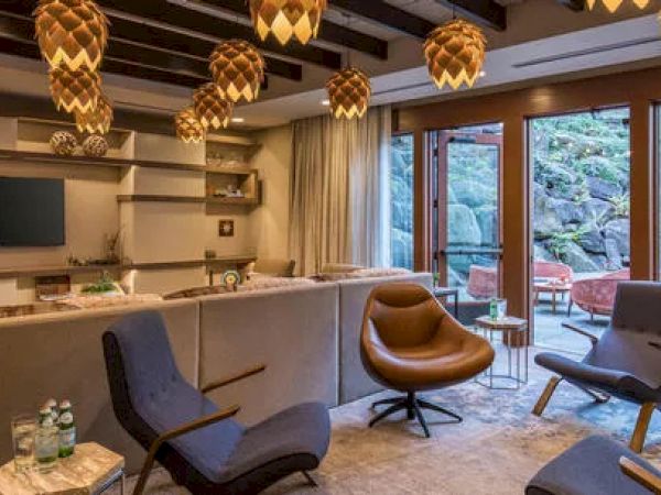 A cozy living room with modern furniture, pendant lights, a TV, and large windows overlooking a rocky outdoor area with seating.