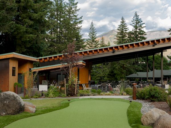 A modern building in a scenic setting with a green lawn and surrounding trees, under a cloudy sky, mountains in the background.