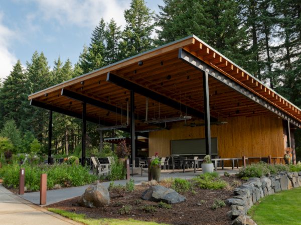 A modern open-air pavilion with a wooden roof is set in a lush, forested area with outdoor seating and landscaped surroundings.