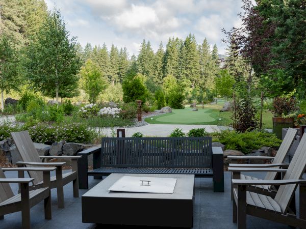 A serene patio setup with wooden chairs and a fire pit, overlooking a lush garden and dense forest in the background.