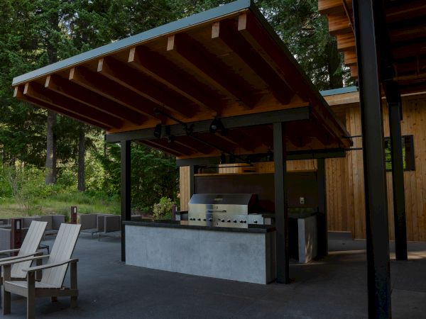 Outdoor kitchen with a wooden canopy, a grill, a countertop, and chairs, surrounded by greenery and a wooden building.