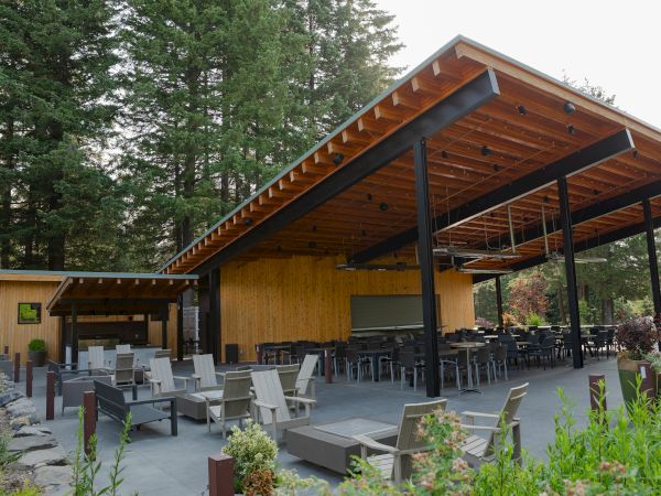 An outdoor seating area with wooden and metal structures, surrounded by lush greenery and trees, featuring chairs and tables.