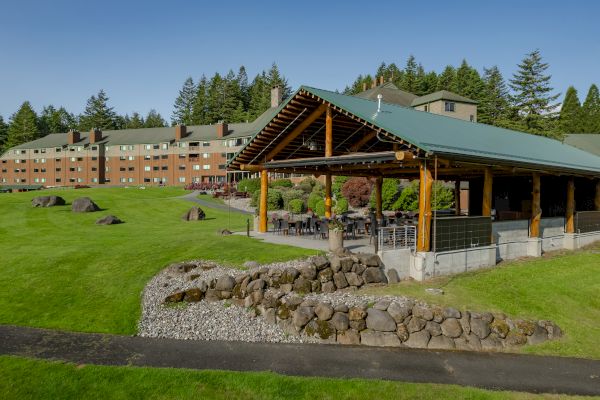 A large building with green roofs is surrounded by a well-maintained lawn and trees, featuring an outdoor seating area under a wooden canopy.