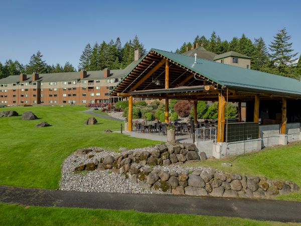 A large building with green roofs is surrounded by a well-maintained lawn and trees, featuring an outdoor seating area under a wooden canopy.