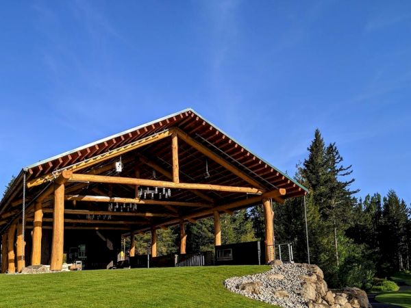A large wooden pavilion with a sloped roof and beams stands on a grassy hill, surrounded by trees under a clear blue sky.