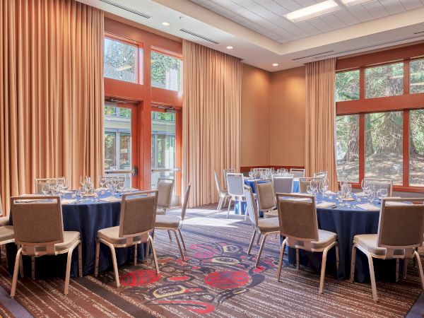 The image shows a banquet room with round tables set with blue tablecloths and chairs, surrounded by large windows and beige curtains.