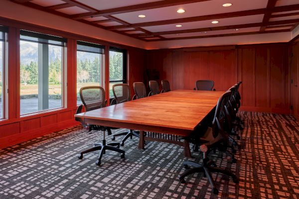 A conference room with a large wooden table, several office chairs, and large windows offering a view of the outdoors.