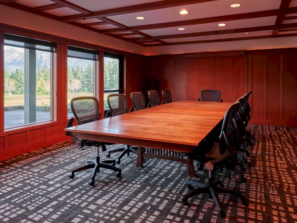 A conference room with a large wooden table, several office chairs, and large windows offering a view of the outdoors.