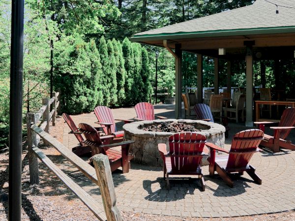 An outdoor patio with Adirondack chairs around a fire pit, surrounded by trees and a wooden fence, under a clear sky.