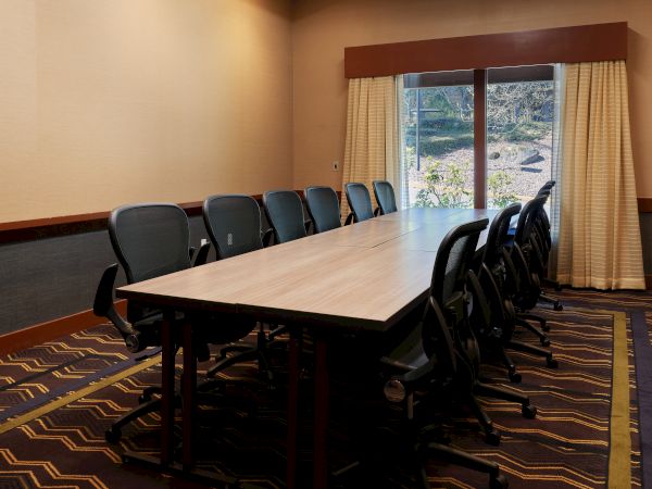 A conference room with a long table, black chairs, patterned carpet, and curtains over a large window in a neutral-toned room.