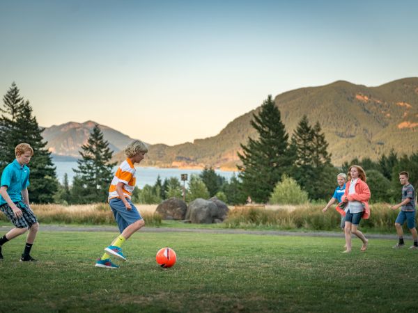 Kids playing soccer in a grassy field with mountains and lake in the background; bright day, everyone moving toward the ball.