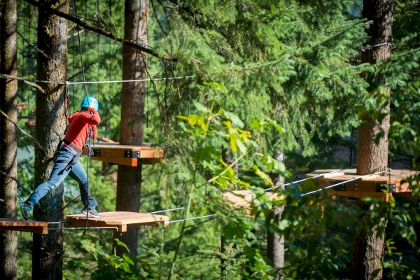 A person in a blue helmet and harness crosses a wooden platform in a forest adventure park.