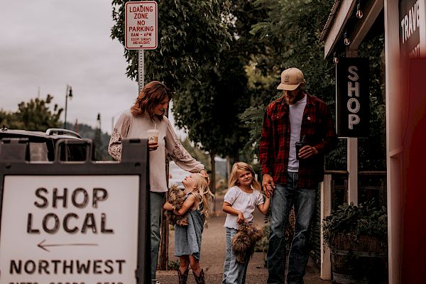 A family walks in a quaint street lined with shops and greenery, passing a "Shop Local Northwest" sign, enjoying a casual day out.