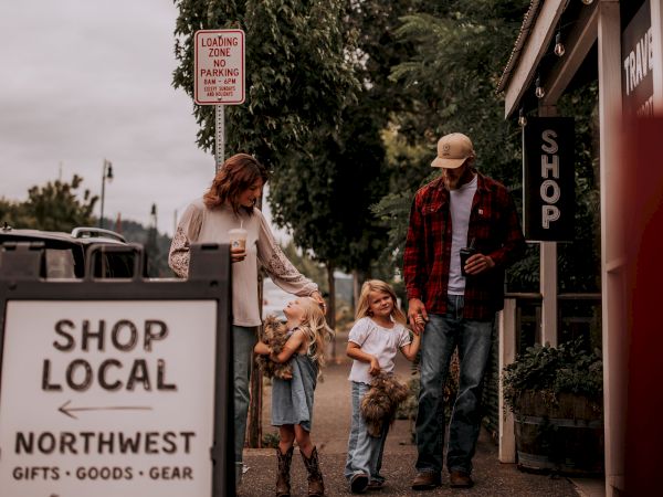 A family walks in a quaint street lined with shops and greenery, passing a "Shop Local Northwest" sign, enjoying a casual day out.
